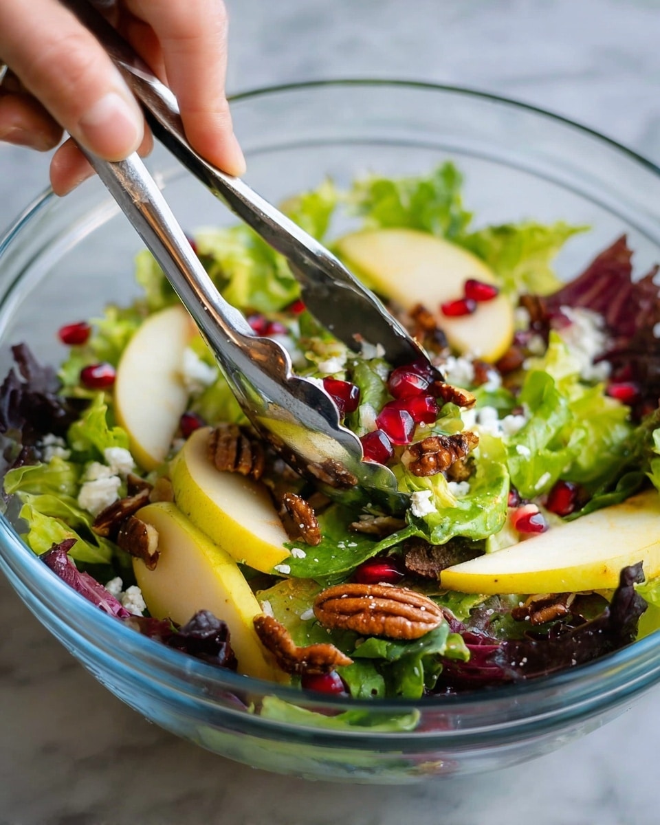 A clear glass bowl holds a fresh salad with multiple layers: the base is green and purple leafy lettuce with soft texture, topped with slices of yellowish apple or pear pieces evenly spread around. Scattered over the leaves are small white crumbled cheese pieces, dark brown pecans, and bright red pomegranate seeds adding color and texture. A woman's hand uses silver metal tongs to lift some salad from the bowl. The bowl sits on a white marbled surface. photo taken with an iphone --ar 4:5 --v 7